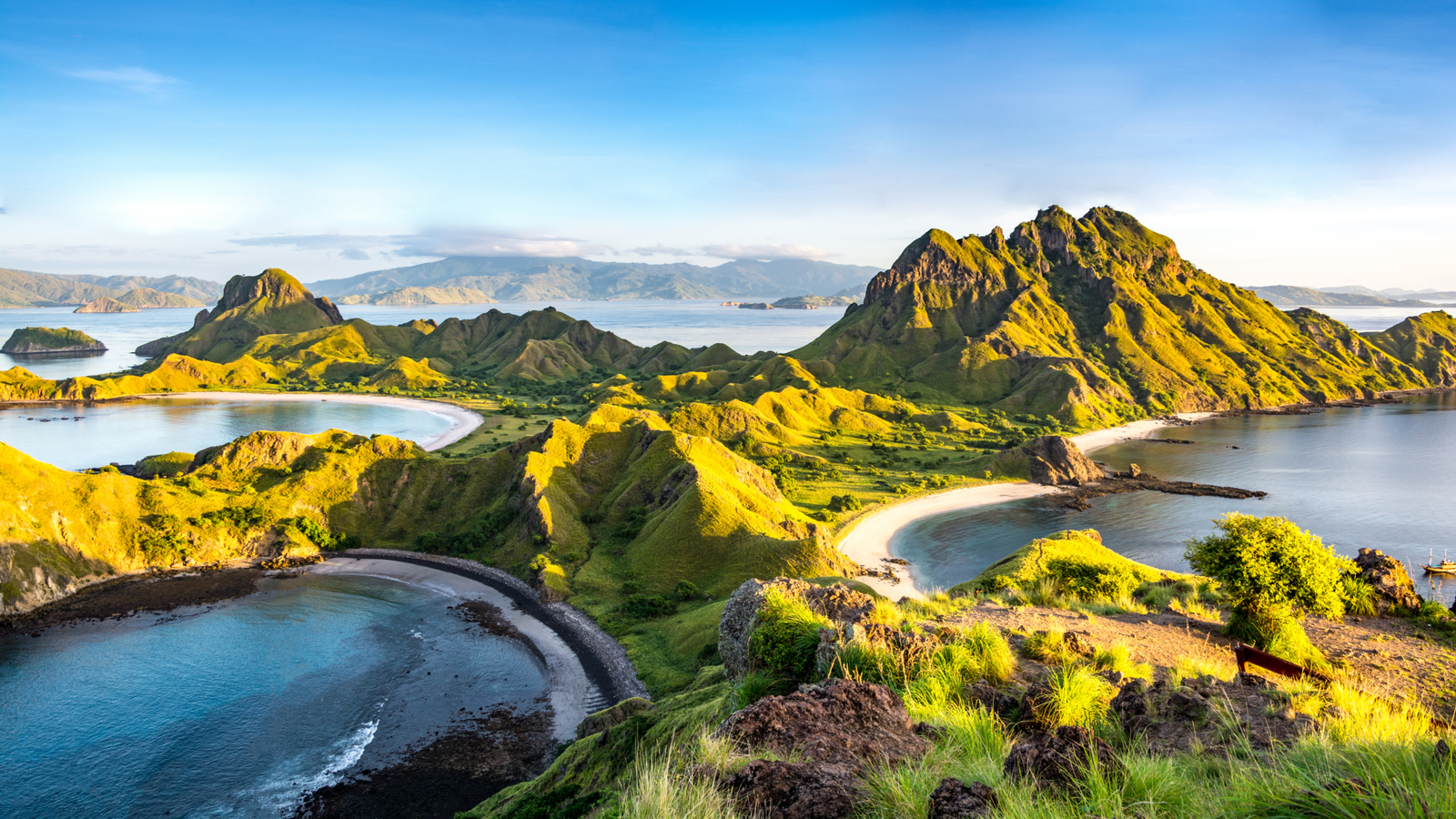 Panoramic view of the Komodo Islands in Indonesia, featuring lush green hills, turquoise waters, pristine white sand beaches, and the dramatic coastal landscape of Komodo National Park under golden morning light.
