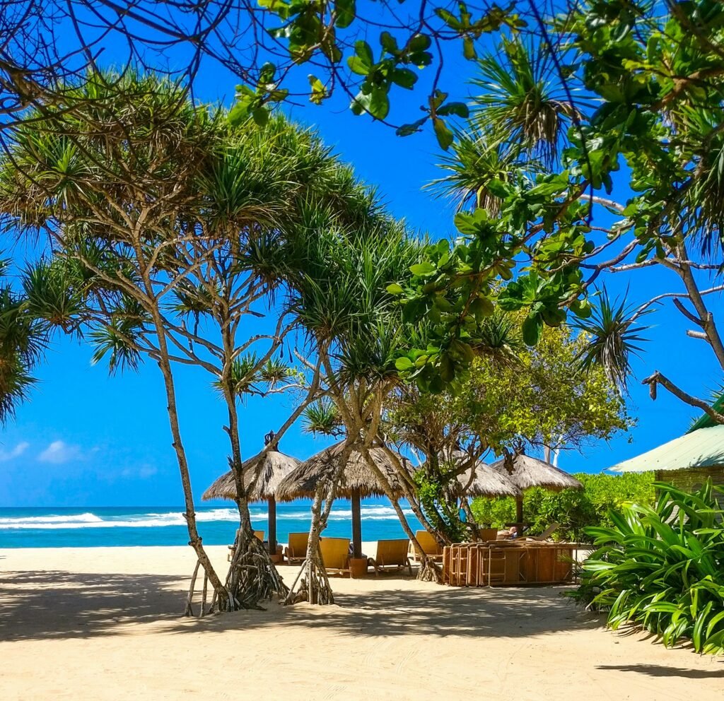 Quiet tropical beach in Nusa Dua, Bali, with white sand, clear blue water and shaded lounge chairs under trees.

