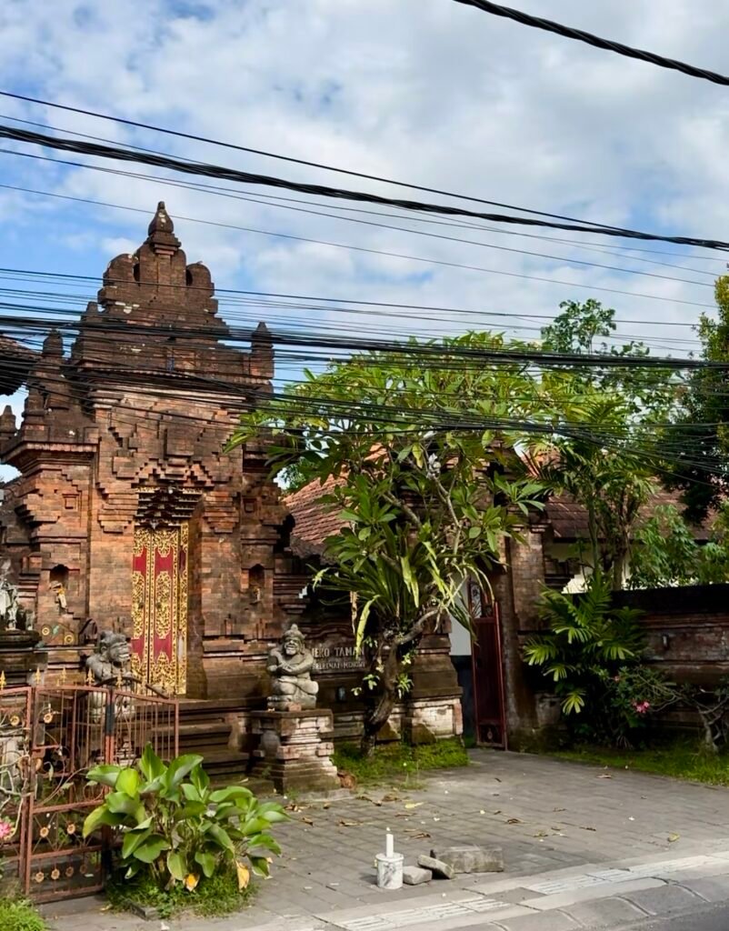 Traditional Balinese entrance gate along a street in Pererenan, Bali, surrounded by tropical plants and overhead power lines.