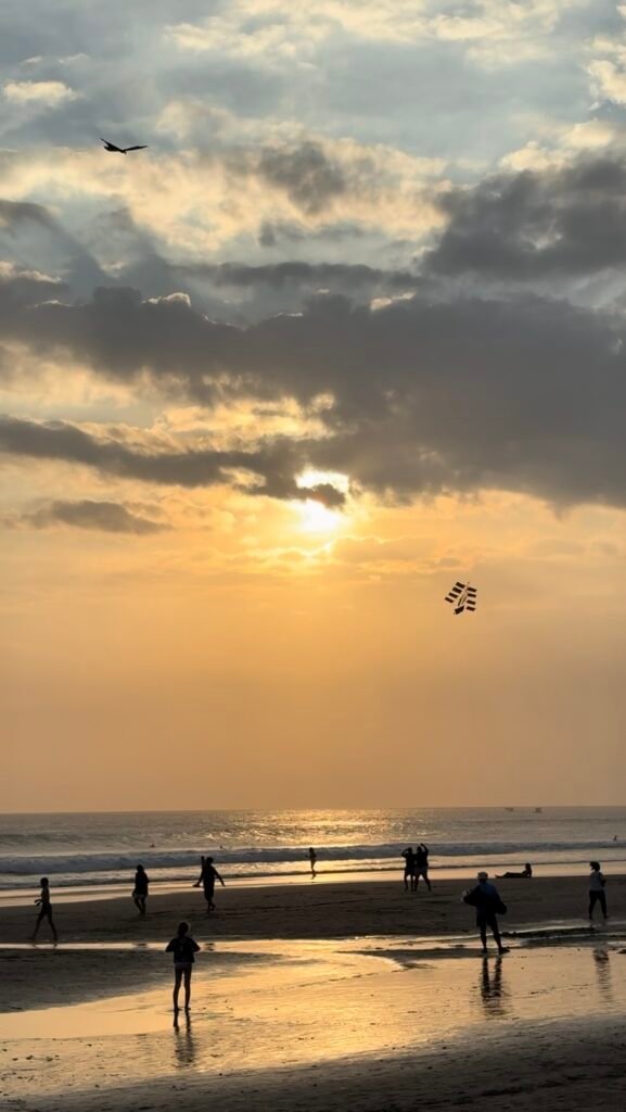Sunset over Seminyak Beach in Bali, with people walking along the shore and a kite flying above the ocean.
